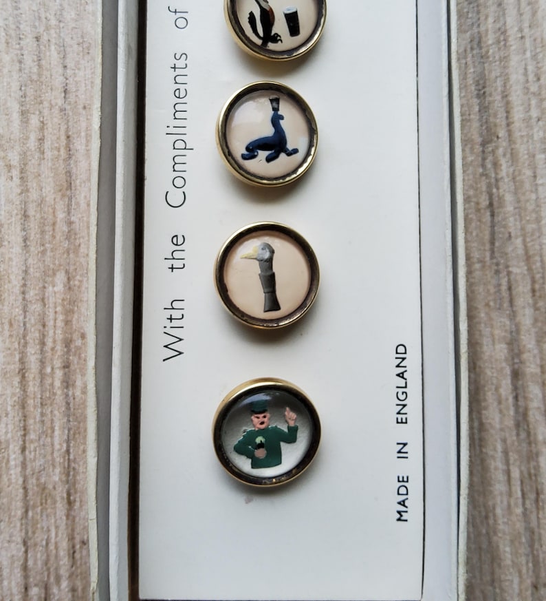 May include: A set of four vintage round buttons with gold-colored rims. Each button features a different whimsical illustration: an eagle, a seal, a duck, and a man. The text "With the Compliments of" and "MADE IN ENGLAND" are also visible.