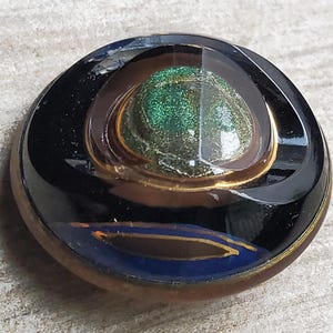Può includere: Bottone rotondo decorativo con un design a strati. Il bottone presenta una parte superiore trasparente a cupola su un elemento centrale verde strutturato. Ha un anello esterno nero, uno strato intermedio marrone e dettagli dorati. Un design blu e oro è visibile nella parte inferiore.