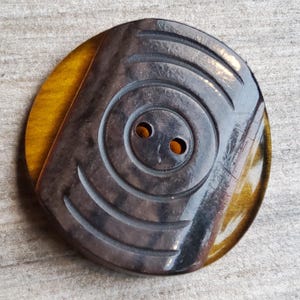 May include: A large, round, two-hole button with a brown and gold tiger eye pattern. The button has a carved design on the front.