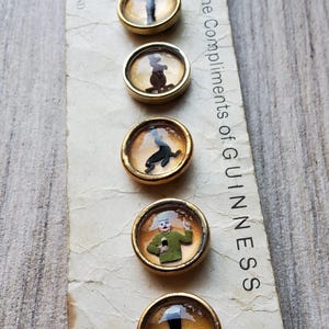 May include: A collection of six vintage Guinness-themed buttons with gold-toned metal rims. Each button features a different whimsical illustration under a clear, domed cover. The paper backing reads "With the Compliments of GUINNESS" and "MADE IN ENGLAND."
