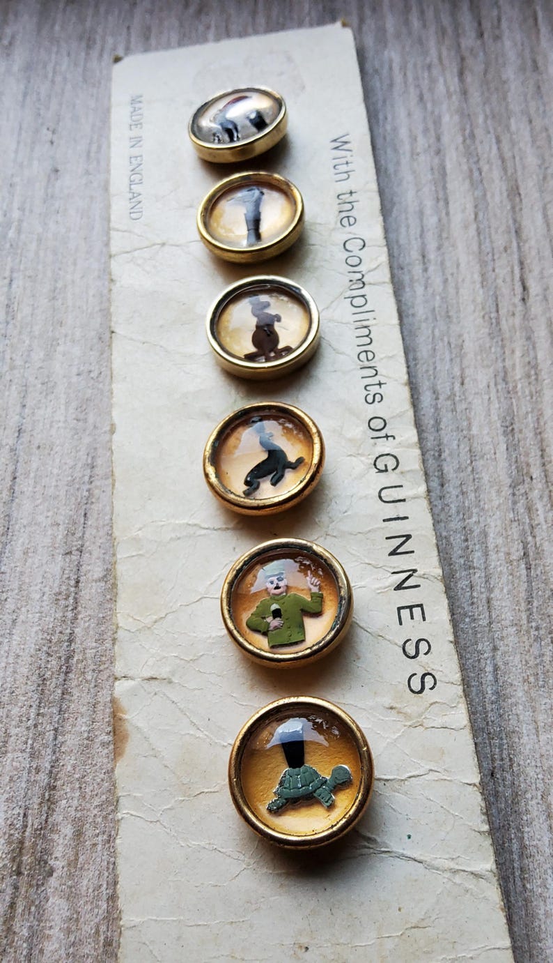 May include: A collection of six vintage Guinness-themed buttons with gold-toned metal rims. Each button features a different whimsical illustration under a clear, domed cover. The paper backing reads "With the Compliments of GUINNESS" and "MADE IN ENGLAND."