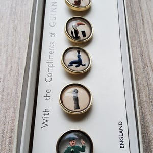 May include: A set of six vintage Guinness-themed buttons with gold-colored rims, each featuring a different whimsical illustration. The buttons are displayed in a white box with the text "With the Compliments of Guinness" and "Made in England".