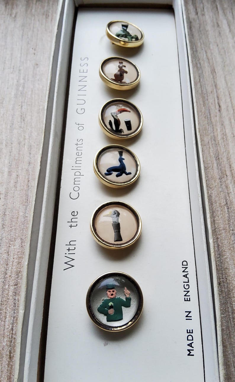 May include: A set of six vintage Guinness-themed buttons with gold-colored rims, each featuring a different whimsical illustration. The buttons are displayed in a white box with the text "With the Compliments of Guinness" and "Made in England".