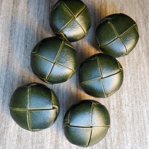 May include: Six olive green, round buttons with a woven leather design. The buttons are arranged on a light brown, textured surface. These decorative buttons are suitable for sewing projects and crafts.