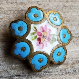 May include: A vintage floral brooch with a gold-toned metal frame. The brooch features a central white enamel design with pink and red flowers. The outer petals are light blue with a gold-toned center.