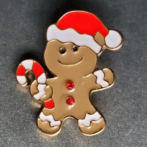 Puede incluir: Un hombre de jengibre con un gorro de Papá Noel rojo y blanco sostiene un bastón de caramelo en su mano derecha. Tiene dos botones rojos en forma de gema en su cuerpo de jengibre marrón.