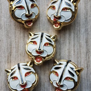 May include: Five gold-coloured tiger head pins with white enamel faces, black stripes, and red eyes and mouths. The pins have a detailed design and are arranged on a wooden surface.