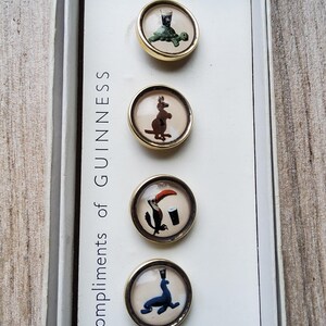 May include: A set of vintage Guinness buttons with various animal illustrations, including a turtle, kangaroo, toucan, and seal. The buttons have gold-colored rims and are displayed in a white box with the text "Compliments of Guinness".