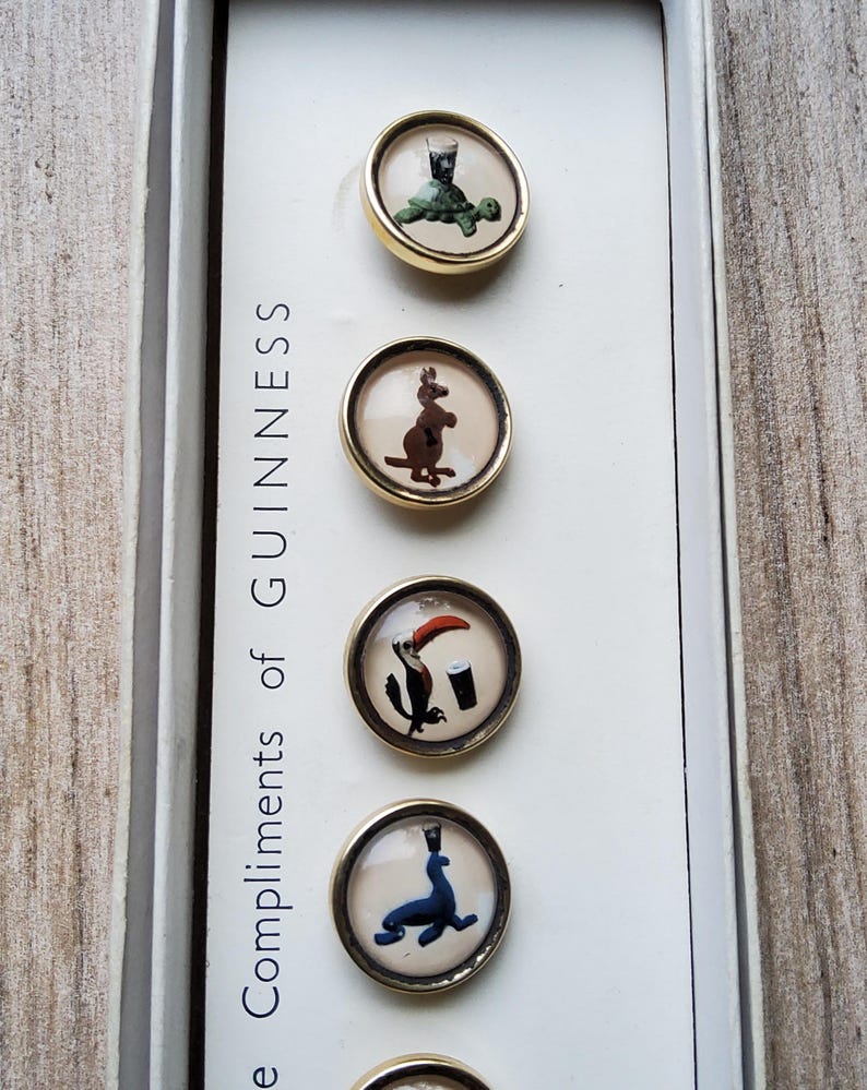 May include: A set of vintage Guinness buttons with various animal illustrations, including a turtle, kangaroo, toucan, and seal. The buttons have gold-colored rims and are displayed in a white box with the text "Compliments of Guinness".