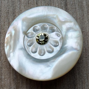 May include: A large, round, iridescent white button with a central floral design. The button features a raised, flower-shaped embellishment with a faceted, olive-green gemstone at its center. The button is made of mother-of-pearl.