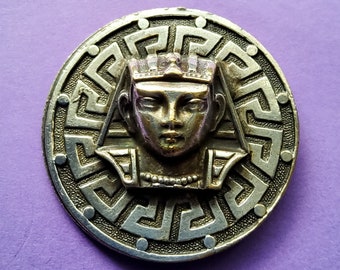 May include: A silver and gold toned metal pin featuring a detailed design of a pharaoh's head wearing a crown, encircled by a Greek key pattern.