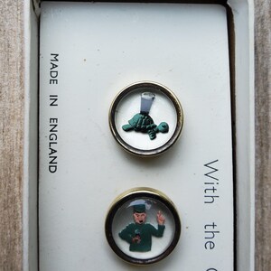 Peut inclure: Deux boutons ronds vintage, dor&eacute;s, avec des d&ocirc;mes en verre transparent. L'un repr&eacute;sente une tortue verte avec un verre de liquide, l'autre une figurine en vert. Le texte "MADE IN ENGLAND" est visible sur le fond blanc.