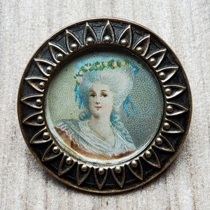 May include: A round, brass-colored locket with a raised, scalloped edge. The locket features a miniature portrait of a woman with a white powdered wig and a blue ribbon. The portrait is set against a blue and white background.