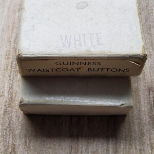 May include: Close-up of two vintage white boxes. The top box has the word "WHITE" printed on it. The bottom box has the words "GUINNESS WAISTCOAT BUTTONS" printed on it. The boxes are slightly worn, suggesting age.