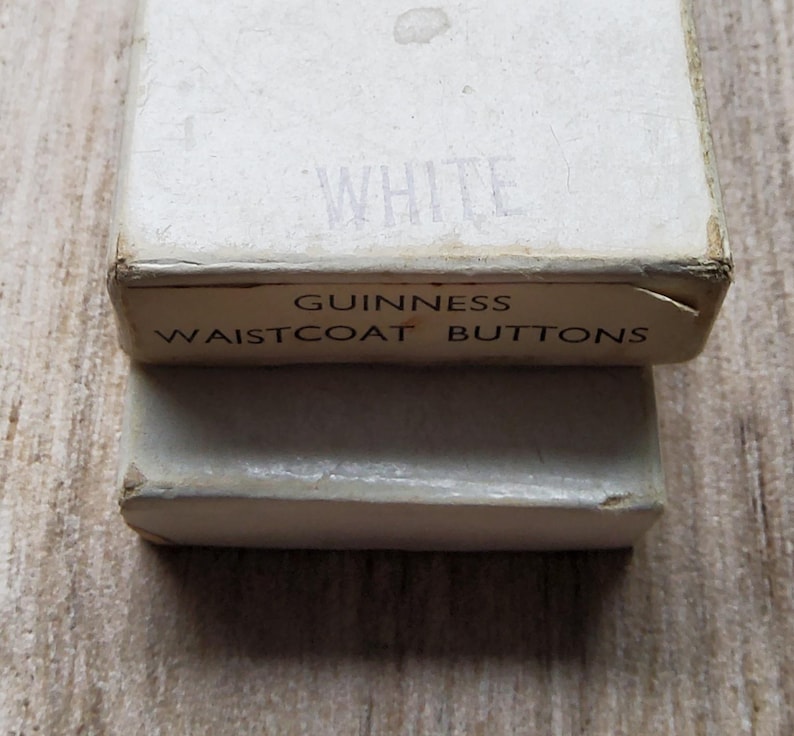 May include: Close-up of two vintage white boxes. The top box has the word "WHITE" printed on it. The bottom box has the words "GUINNESS WAISTCOAT BUTTONS" printed on it. The boxes are slightly worn, suggesting age.