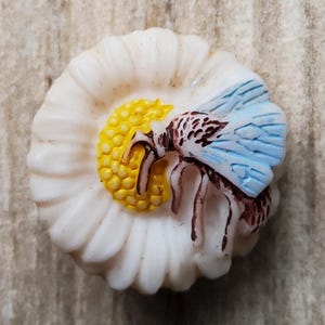 May include: A white daisy-shaped decorative object with a yellow center and a bee with blue wings. The bee has brown and white markings. The daisy petals are white and curved. The object is on a light brown surface.