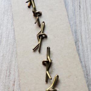 May include: Close-up of several small, brass-colored cotter pins arranged on a light brown surface. The pins are bent into a looped shape, with a split end for securing. The background is a light wood grain.