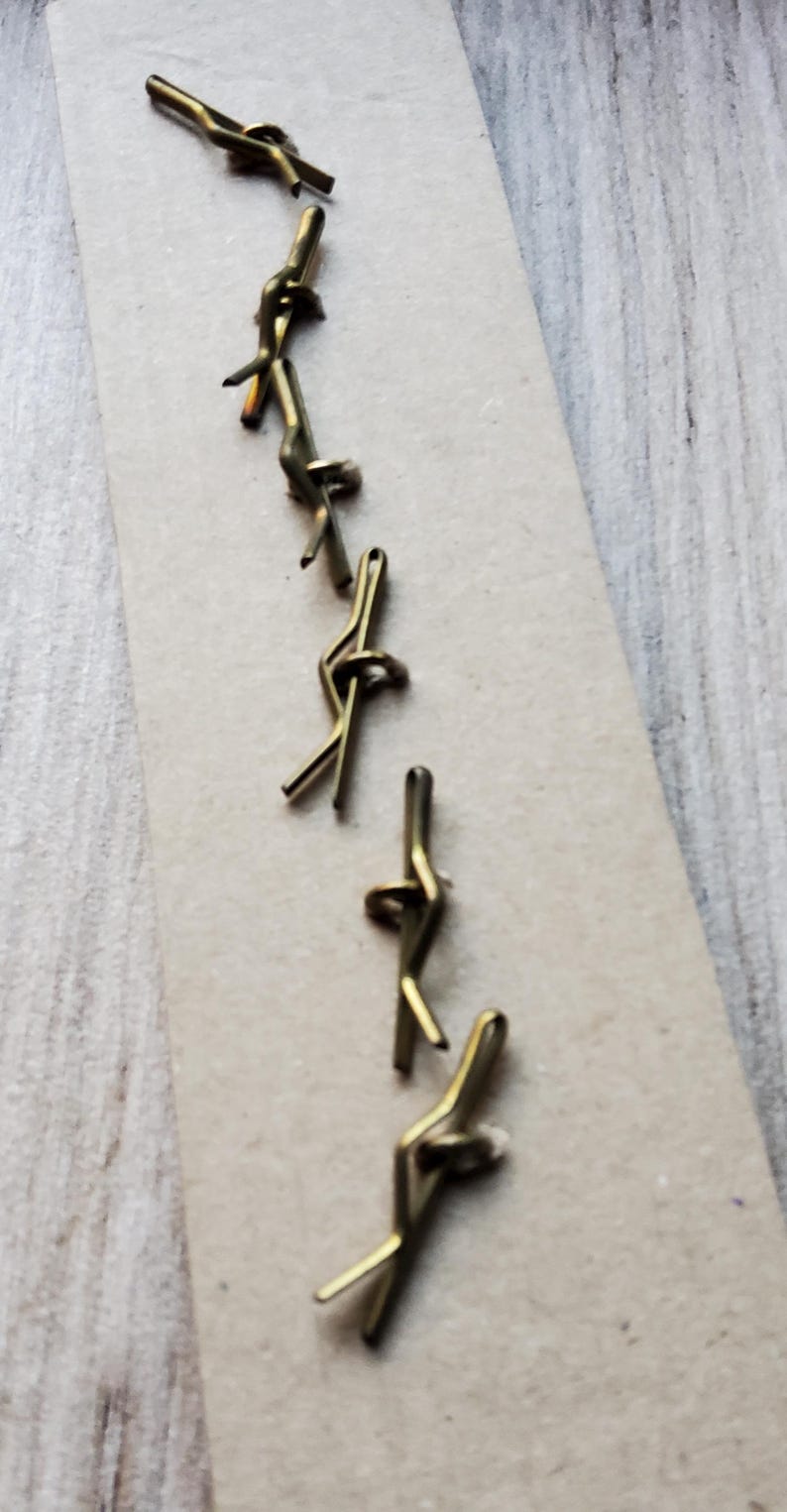 May include: Close-up of several small, brass-colored cotter pins arranged on a light brown surface. The pins are bent into a looped shape, with a split end for securing. The background is a light wood grain.
