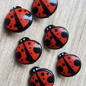 Peut inclure: Un ensemble de cinq boutons ronds en forme de coccinelles, chacun présentant un corps rouge vif avec des points noirs, une tête noire avec deux petits points blancs et une finition brillante. Les boutons sont disposés sur une surface claire.