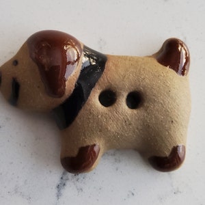 May include: A ceramic button shaped like a brown dog with black accents. The dog has two holes for sewing.