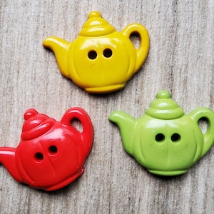 Puede incluir: Tres botones con forma de tetera en amarillo, rojo y verde. Cada botón tiene dos agujeros y un asa. Los botones están dispuestos sobre una superficie de madera clara, mostrando sus colores brillantes y su diseño juguetón.
