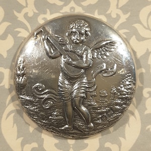 May include: A silver-toned round button with a raised design of an angel playing a musical instrument. The angel has wings and is surrounded by foliage.