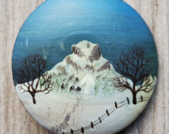 May include: A hand-painted wooden ornament depicting a snowy mountain scene with two bare trees and a white fence. The sky is a light blue color.