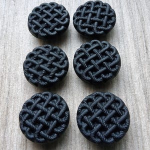 May include: Six black, round buttons featuring a woven rope pattern. The buttons have a textured surface and are arranged in two rows of three on a light wood grain background.