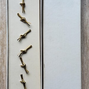 Peut inclure: Bo&icirc;te ouverte contenant six pinces &agrave; cravate dor&eacute;es en forme de n&oelig;ud. Les pinces sont dispos&eacute;es verticalement sur un fond cr&egrave;me. La bo&icirc;te blanche porte le mot "WHITE" imprim&eacute; sur le couvercle.