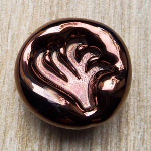 May include: A round, metallic button with a raised, stylized floral design. The button has a glossy, copper-coloured finish with a reflective surface. The floral design is a lighter, pinkish hue.
