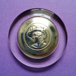 May include: A silver button with a raised image of a woman wearing a hat. The button is encased in a clear plastic dome.