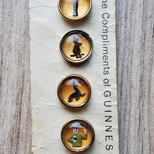 May include: A vintage card featuring six round, gold-toned pins with various Guinness-themed illustrations. The card reads "With the Compliments of GUINNESS" and "MADE IN ENGLAND". The pins depict a harp, a pint, a dog, a seal, a man, and a turtle.