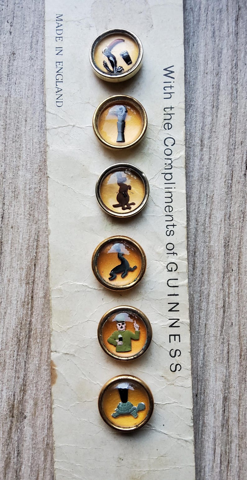 May include: A vintage card featuring six round, gold-toned pins with various Guinness-themed illustrations. The card reads "With the Compliments of GUINNESS" and "MADE IN ENGLAND". The pins depict a harp, a pint, a dog, a seal, a man, and a turtle.