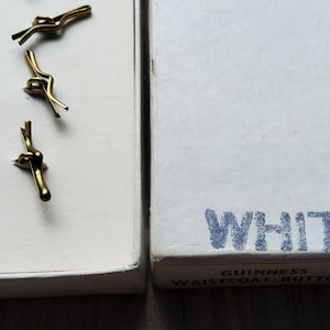 Peut inclure: Trois boutons de gilet dor&eacute;s dans une bo&icirc;te blanche. La bo&icirc;te &agrave; c&ocirc;t&eacute; est blanche avec le mot "WHITE" en lettres bleues. La bo&icirc;te est &eacute;tiquet&eacute;e "Guinness Waistcoat Buttons".