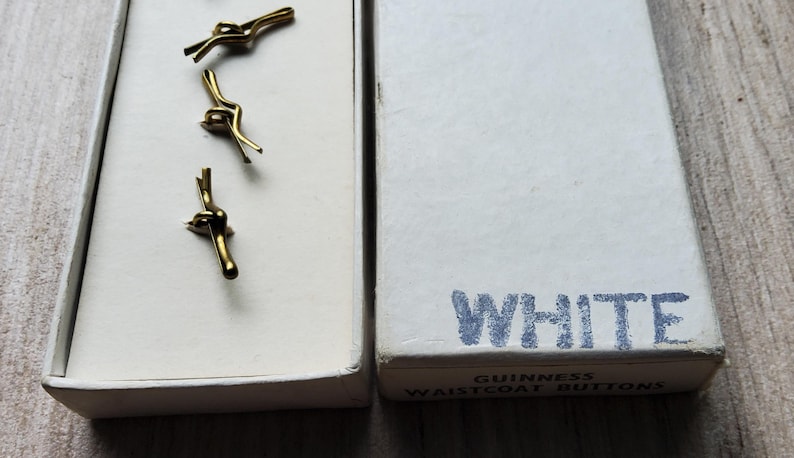 Peut inclure: Trois boutons de gilet dor&eacute;s dans une bo&icirc;te blanche. La bo&icirc;te &agrave; c&ocirc;t&eacute; est blanche avec le mot "WHITE" en lettres bleues. La bo&icirc;te est &eacute;tiquet&eacute;e "Guinness Waistcoat Buttons".