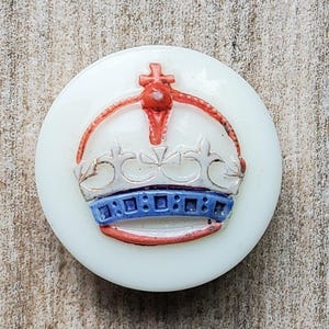 Pode incluir: Um botão redondo branco sujo com um design detalhado de coroa. A coroa apresenta detalhes vermelhos, azuis e prateados, com uma cruz no topo. O botão é provavelmente feito de plástico ou resina, adequado para costura ou artesanato.