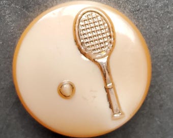 Può includere: Un bottone bianco con un'immagine in rilievo di una racchetta da tennis e una palla. La racchetta è argentata con un manico nero e la palla è bianca.