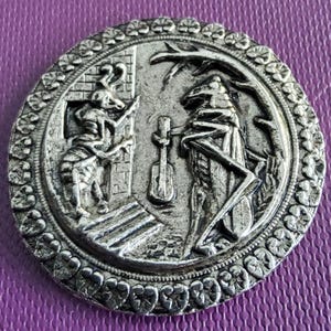 May include: A silver-toned, circular button with a detailed relief design. The central image depicts two insect-like figures, one playing a stringed instrument. The border features a repeating floral pattern. The button has an antique or vintage aesthetic.