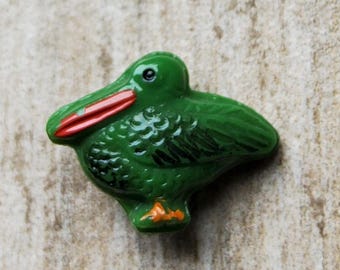 Puede incluir: Pequeña figura de pájaro verde con pico rojo y patas naranjas. El pájaro tiene detalles negros en sus alas y cuerpo. La figura es de material brillante y está sobre un fondo neutro.