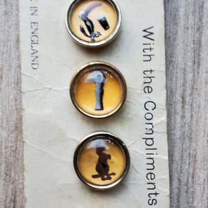 May include: Set of four vintage round buttons with gold-toned metal rims. Each button features a different black silhouette design on a yellow background. The text "Made in England" is visible. The text "With the Compliments of G" is also visible.