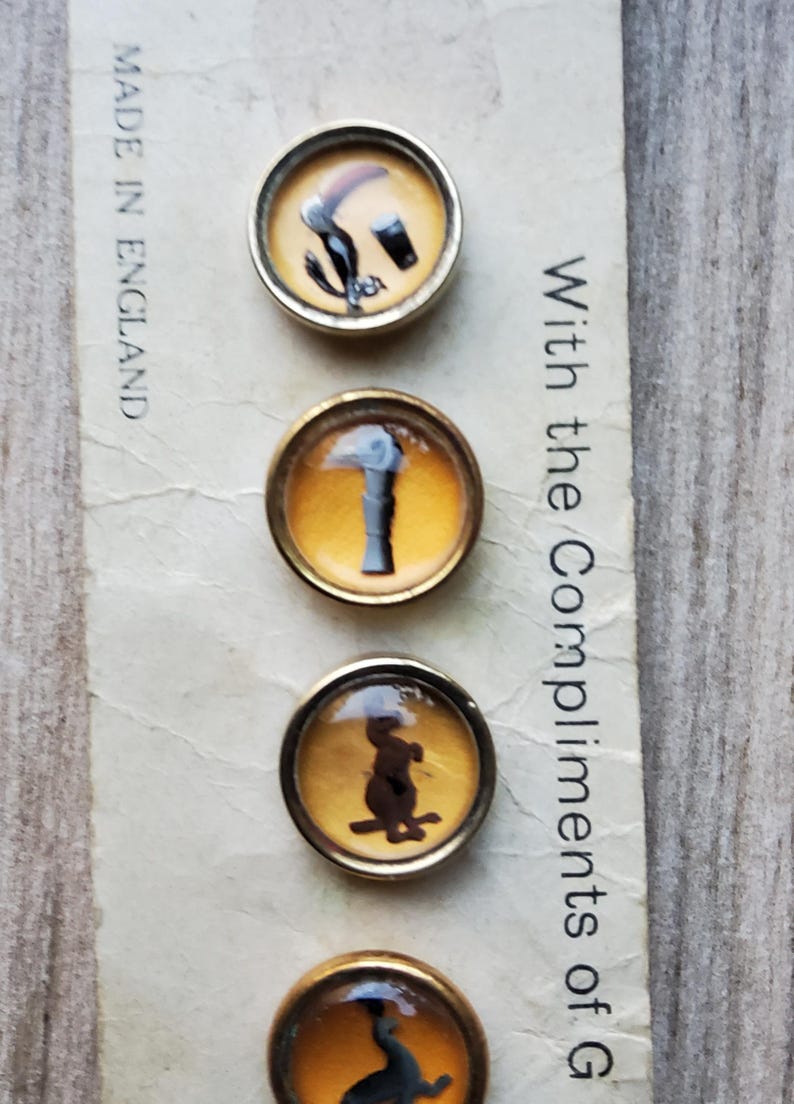 May include: Set of four vintage round buttons with gold-toned metal rims. Each button features a different black silhouette design on a yellow background. The text "Made in England" is visible. The text "With the Compliments of G" is also visible.