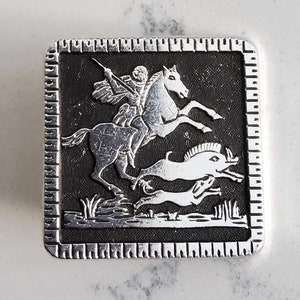 May include: A silver-toned metal square-shaped pin with a black background. The pin depicts a rider on horseback hunting a boar and a smaller dog. The pin has a decorative border.
