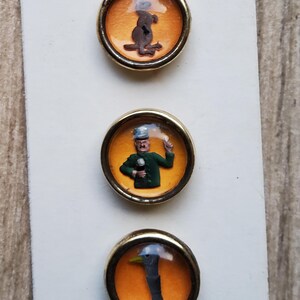 May include: Three round, gold-rimmed buttons with orange backgrounds and clear glass tops. The buttons feature images of a kangaroo, a man in a green uniform, and a bird's head.