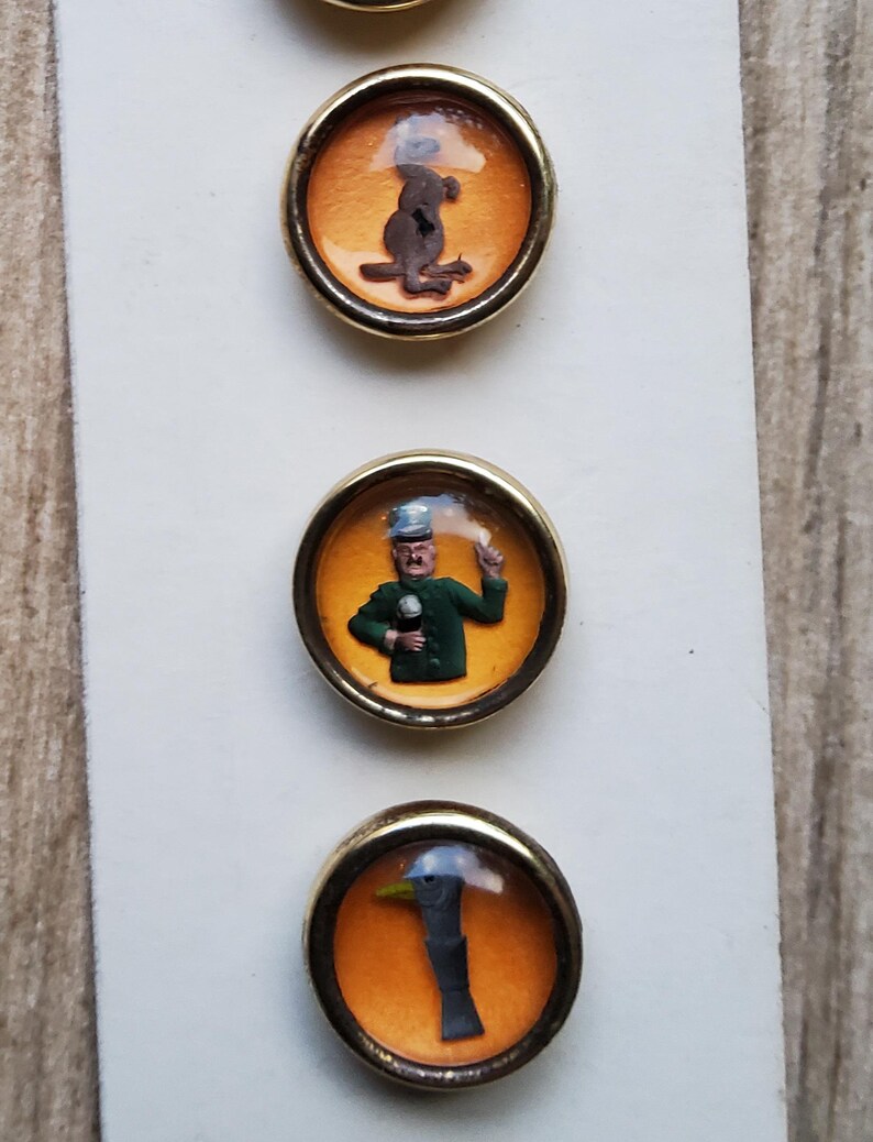 May include: Three round, gold-rimmed buttons with orange backgrounds and clear glass tops. The buttons feature images of a kangaroo, a man in a green uniform, and a bird's head.