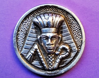 May include: A silver-toned metal pendant featuring a detailed relief of an Egyptian pharaoh wearing a headdress. The pharaoh is holding a staff and a crook in his hands.