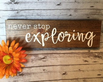 May include: A rectangular wooden sign with the words "never stop exploring" in white lettering. An orange and yellow flower is in the lower left corner. The sign is on a wooden background.
