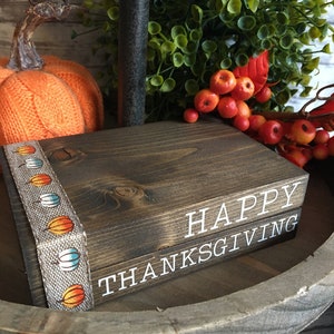 May include: A wooden block sign with the words "HAPPY THANKSGIVING" in white lettering. The sign is decorated with a ribbon featuring colorful pumpkin designs. An orange pumpkin and fall foliage are in the background.