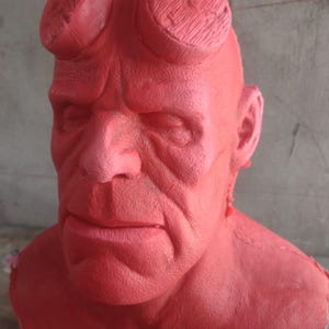Könnte beinhalten: Rote Büste einer Figur mit ernstem Gesichtsausdruck. Die Figur hat eine große Stirn, markante Augenbrauen und zwei große, abgerundete Hörner. Die Haut ist strukturiert, und das Gesamtbild deutet auf einen geformten oder geformten Gegenstand hin.
