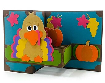 May include: A pop-up card featuring a brown turkey with a yellow, purple, and orange feathered tail. The turkey is sitting on a green and brown platform with two orange pumpkins. The card has a blue background with pink and yellow leaves.