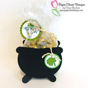 Może przedstawiać: Czarny papierowy kocioł w kształcie kotła z uchwytem ze złotego sznurka. Kocioł jest wypełniony przezroczystym workiem złotych czekoladowych monet. Do uchwytu przymocowana jest zielona etykieta z koniczyną. Etykieta głosi "Wesołych Świąt Świętego Patryka!"